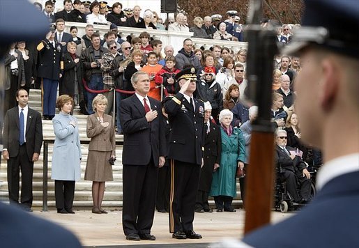 Placing his hand over his heart, President George W. Bush participates in the Wreath Laying Ceremony at the Tomb of the Unknowns in Arlington Cemetery on Veterans Day Nov. 11, 2003. Laura Bush is pictured standing behind the President. White House photo by Paul Morse.