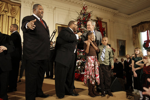 President George W. Bush is joined by two children on stage in the East Room of the White House as he thanks members of the musical group Sweet Heaven Kings, Monday, Dec. 8, 2008, during the Children's Holiday Reception and Performance. White House photo by Eric Draper