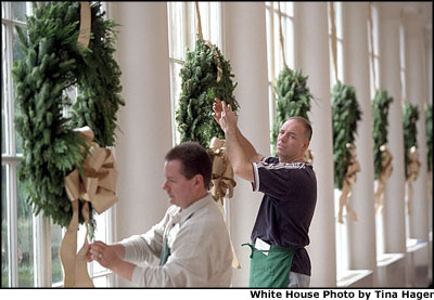Volunteers Andy Wentz (left) and Aaron Vincelette hang wreaths along the windows of the East Colonnade. Overlooking the Jacqueline Kennedy garden, the walkway is adorned with wreaths hung with gold ribbon. White House photo by Tina Hager.