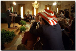 President George W. Bush addresses the audience in the East Room as actor Rob Schneider, decked out in a patriotic cowboy hat, takes a photograph Nov. 2. White House photo by Eric Draper.