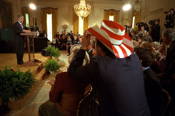 President George W. Bush addresses the audience in the East Room as actor Rob Schneider, decked out in a patriotic cowboy hat, takes a photograph Nov. 2. White House photo by Eric Draper.