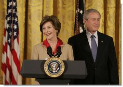 President George W. Bush listens on as Mrs. Laura Bush welcomes women leaders to the East Room for a celebration Tuesday, March 7, 2006, of International Women's Day. Mrs. Bush said, "I've been privileged to meet thousands of women from many nations, and I believe that women everywhere share the same dreams -- to be educated, to live in peace, to enjoy good health, to be prosperous, and to be heard."  White House photo by Kimberlee Hewitt