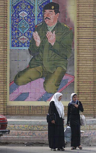 Photo of women standing in front of a mural of Saddam Hussein in prayer