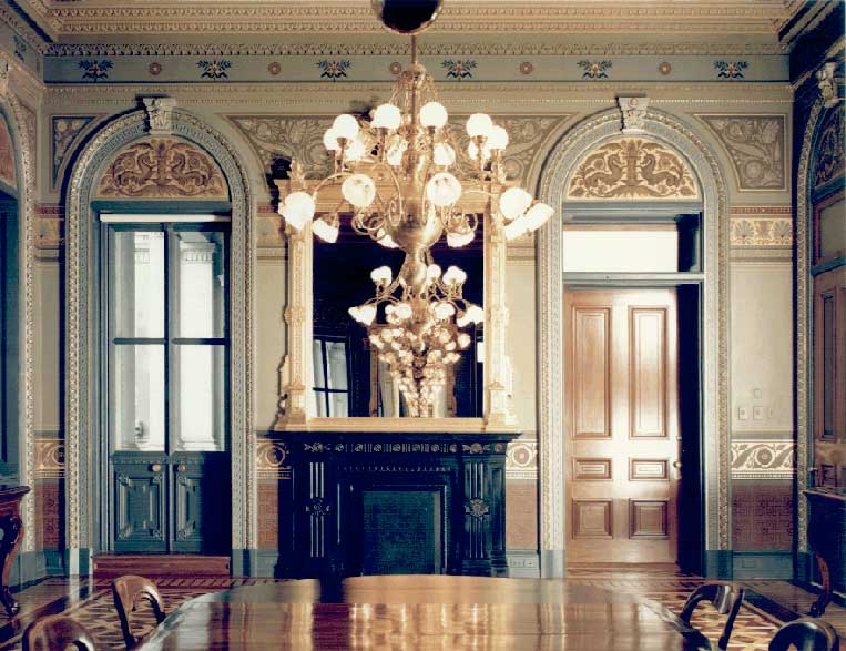 Color photo of the Vice President's Ceremonial Office. Pictured is an elegant conference table, a chandelier, and a two windows topped with arches and ornate stenciling.